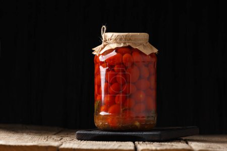 Photo for Canned cherry tomatoes in a closed glass jar with a craft lid on a wooden surface, dark background, space for text. - Royalty Free Image