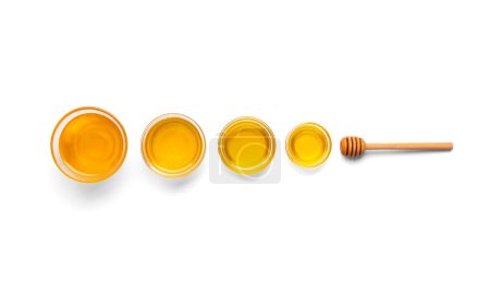 Foto de Composición creativa de miel en cuencos de vidrio de diferentes tamaños y una cacerola de madera en una vista superior de fondo blanco. Miel orgánica y cuchara de miel en aislamiento. - Imagen libre de derechos
