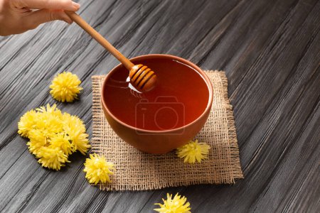 Foto de Miel en un tazón de barro, cazo y flores amarillas sobre un textil y un fondo de madera oscura. Un palo para la miel se encuentra en un tazón de cerámica con la miel de cerca. - Imagen libre de derechos