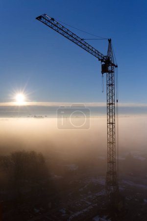 Téléchargez les photos : Grue de chantier dans le brouillard le matin. Lever de soleil, brouillard matinal, chantier, grue à tour. Mystique matin brumeux dans la ville à l'aube. Paysage urbain. - en image libre de droit