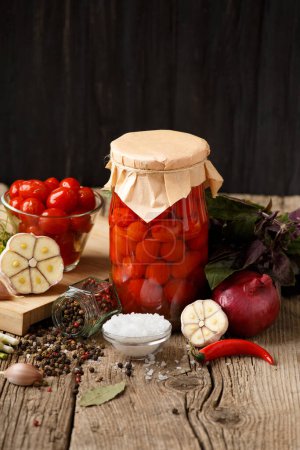 Téléchargez les photos : Tomates cerises en conserve dans un bocal fermé et un bol en verre, épices et herbes pour la marinade sur un fond de bois rustique. - en image libre de droit