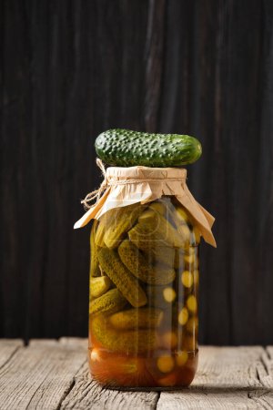 Photo for Pickled cucumbers in a closed glass jar with a craft lid, one fresh gherkin lies on a jar, wooden surface, dark background, copy space. - Royalty Free Image