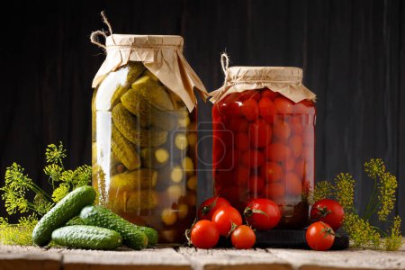 Pepinos enlatados pepinillos y tomates cherry en frascos, verduras frescas para su conservación sobre un fondo de madera oscura.