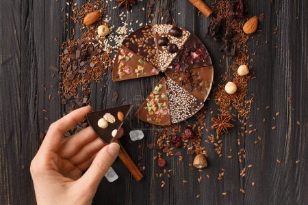 Photo for Chocolate pizza with nuts, dry berries, sesame seeds and ingredients on a dark wooden background top view. The hand holds a piece of sweet chocolate pizza. - Royalty Free Image