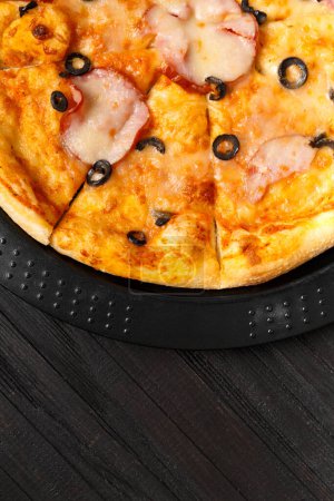 Photo for Pizza with jerky, olives, cheese and herbs on a black round baking dish, top view. Traditional Italian food. - Royalty Free Image