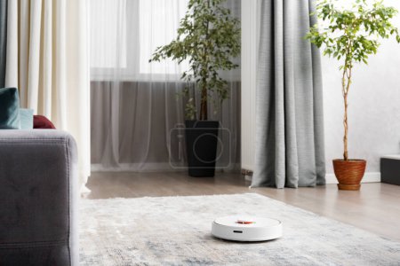 Téléchargez les photos : Aspirateur robot intelligent dans un salon moderne. Le concept d'une maison intelligente, nettoyage sans fil par un robot avec un aspirateur de toutes les surfaces. - en image libre de droit