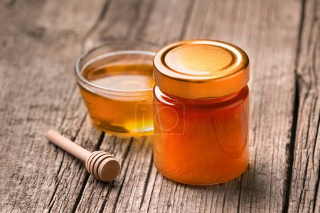 Organic honey in a closed glass jar and in a transparent glass bowl, wooden dipper on a rustic wooden background, close-up.