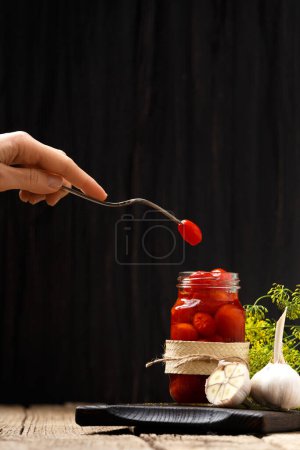 Photo for Pickled cherry tomatoes in an open jar on a dark wooden board, one tomato on a vintage fork in hand, dark background, close-up. - Royalty Free Image
