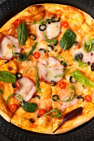 Foto de Pizza con jerky, aceitunas, queso y hierbas en un plato negro redondo para hornear, vista superior. Comida tradicional italiana. - Imagen libre de derechos