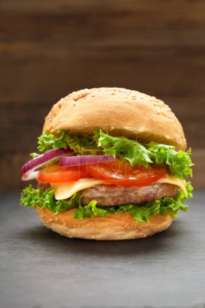Foto de Hamburguesa apetitosa con jugosa chuleta de res, queso, tomates, lechuga, cebolla en un plato de piedra negra sobre un fondo de madera oscura. Comida rápida, hamburguesa casera. - Imagen libre de derechos