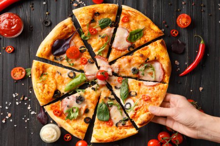 Photo for Hands take slices of cut pizza on a dark wooden background top view. Pizza with salami, jerky, sauces, spices, fresh cherry tomatoes on a black background. Women's hands share a pizza. - Royalty Free Image