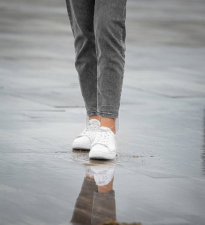 Photo for Legs of a girl in jeans and white sneakers walk in rainy weather on a street in the city, raindrops, water splashes, reflection in puddles - Royalty Free Image