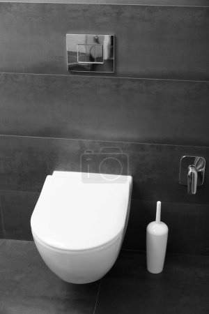 Foto de Un moderno inodoro blanco montado en la pared con una tapa cerrada y un botón de color cromo brillante contra una pared negra. Tazón de inodoro en el interior del baño. - Imagen libre de derechos