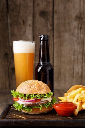 Foto de Hamburguesa, cerveza en una botella y en una taza de vidrio, patatas fritas, salsa sobre un fondo de madera, concepto de comida rápida. - Imagen libre de derechos