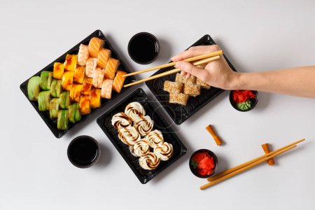 Téléchargez les photos : Un ensemble de rouleaux de sushi lumineux avec des crevettes et du saumon dans des emballages en plastique sur un fond blanc, la main d'une femme prend le rouleau avec des baguettes chinoises, vue du dessus. Livraison de sushi - en image libre de droit
