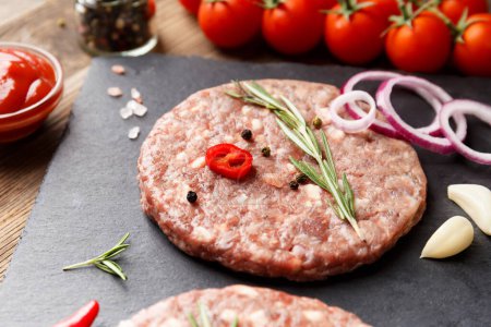 Photo for Raw fresh ground beef burger patties, rosemary, vegetables and spices on a black stone plate on a wooden background. Ingredients for a hamburger. - Royalty Free Image