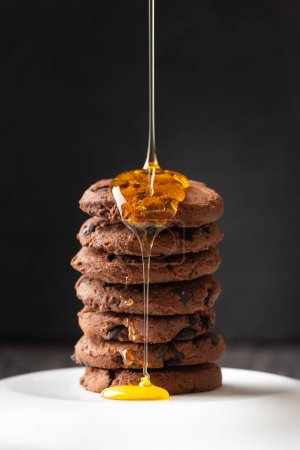 Téléchargez les photos : Honey pours on homemade chocolate chip cookies on a dark background close-up. Composition of craft cookies with pieces of chocolate and honey pouring over it. Dessert. - en image libre de droit