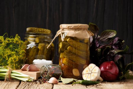 Photo for Pickled cucumbers in closed and open jars, onion, garlic, dill, parsley, basil on a dark wooden background. Homemade canned gherkins , spices and herbs for marinade. - Royalty Free Image