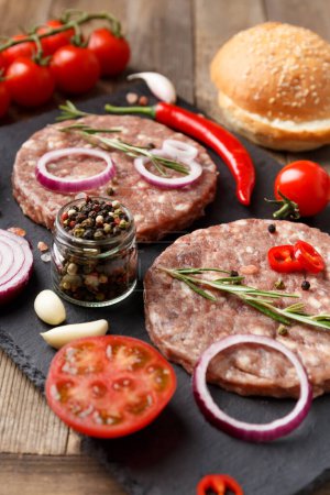 Photo for Raw fresh ground beef burger patties, rosemary, vegetables and spices on a black stone plate on a wooden background. Ingredients for a hamburger. - Royalty Free Image