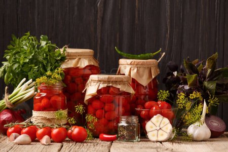 Téléchargez les photos : Tomates cerises en conserve dans des pots fermés et ouverts, épices et herbes pour la marinade sur un fond de bois rustique. - en image libre de droit