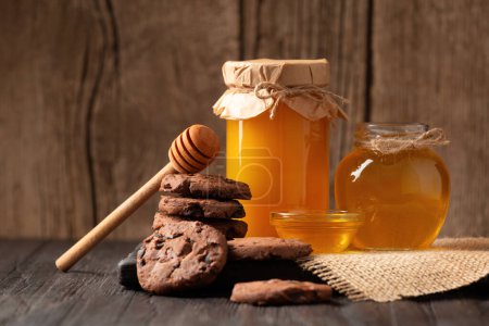 Photo for Honey in jars and in a bowl, dipper and homemade cookies with chocolate chips on a wooden background. Homemade cakes and honey for breakfast. Composition of craft chocolate cookies and honey. - Royalty Free Image