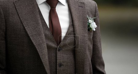 Foto de El novio lleva un elegante traje de novia en tonos marrones, una corbata y un boutonniere en el ojal. Moda de boda elegante. - Imagen libre de derechos