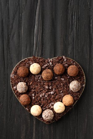 Photo for White, milk and dark chocolate truffles and chocolate chips in a heart-shaped box on a dark wooden background, top view. Valentine's Day. - Royalty Free Image