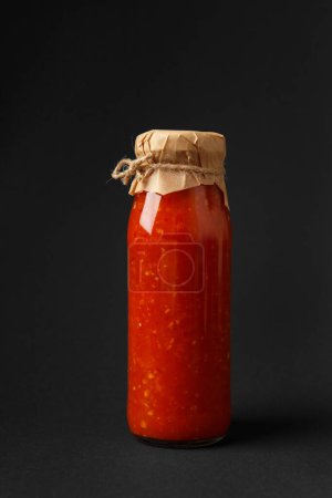 Téléchargez les photos : Sauce piquante à base de piments, paprika, tomates et ail dans un bocal en verre, isolé sur un fond noir. Sauce blanche, adjika. - en image libre de droit