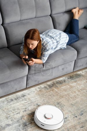 Foto de La chica se encuentra en el sofá de la sala de estar y controla la aspiradora robot con un teléfono inteligente. Casa Inteligente. El concepto de electrodomésticos inteligentes, tareas domésticas y tecnología. - Imagen libre de derechos