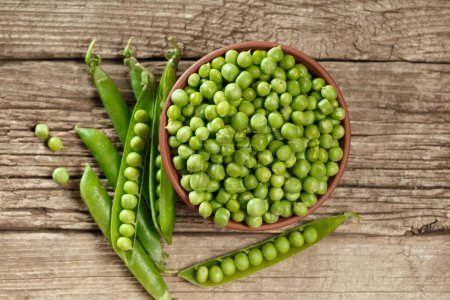 Photo for Peeled green peas in a clay bowl, sweet organic green peas in closed and open pods and scattered grains on an aged wooden background, top view. vegetable protein. - Royalty Free Image