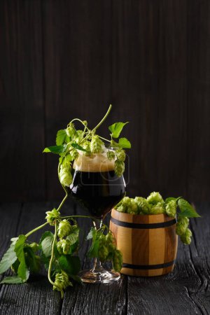 Photo for Glass mug of dark beer, branches of hops and a wooden barrel with hops on a dark wooden background. Brewing traditions. Oktoberfest. Beer festival. St.Patrick 's Day - Royalty Free Image