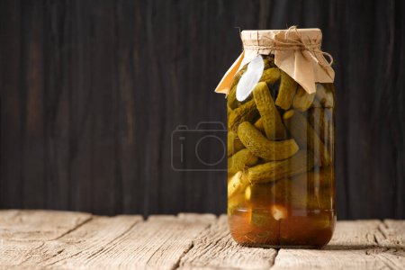 Photo for Pickled cucumbers in a closed glass jar on a wooden surface with space for text. Homemade canned cucumbers in a bottle, copy space. - Royalty Free Image