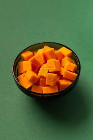 Foto de Calabaza naranja dulce cortada en cubos en un plato negro sobre un fondo verde. Plantilla culinaria para platos de calabaza. Alimentación saludable. Trozos de calabaza. - Imagen libre de derechos