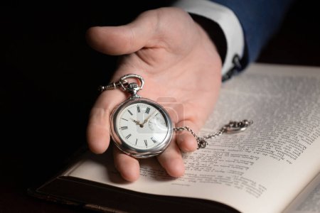 Téléchargez les photos : Une main tient une montre de poche vintage sur le fond d'un vieux livre. Concept de temps rapide, passé, sagesse, expérience - en image libre de droit