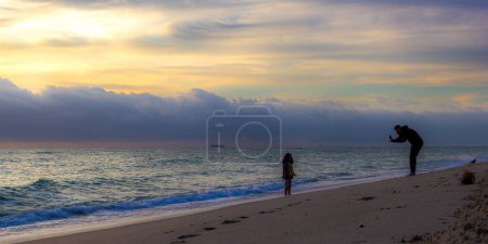 Photo for Father taking a picture of his doughter, sunset on the beach. - Royalty Free Image
