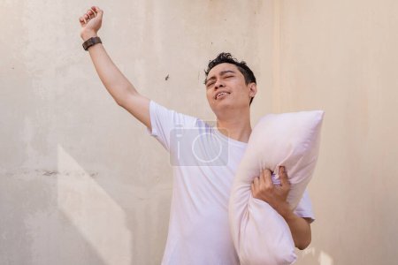 Foto de Los hombres indonesios se sienten perezosos y somnolientos sostienen la almohada con una camiseta blanca. La foto es conveniente utilizar para la publicidad de la expresión del hombre y el estilo de vida de la manera. - Imagen libre de derechos