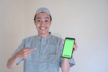 Téléchargez les photos : Homme musulman asiatique heureux, souriant lorsque montrer le smartphone avec écran vert lors de la célébration du Ramadan. La photo est appropriée pour l'affiche Ramadhan et les médias de contenu musulman. - en image libre de droit