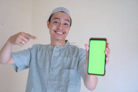 Téléchargez les photos : Homme musulman asiatique heureux, souriant lorsque montrer le smartphone avec écran vert lors de la célébration du Ramadan. La photo est appropriée pour l'affiche Ramadhan et les médias de contenu musulman. - en image libre de droit