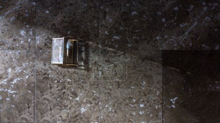 Foto de Wall covered with marble tiles with a muted wall light on which a ray of sunlight from outside falls. Copyspace. Panoramic format. - Imagen libre de derechos
