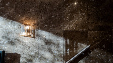Foto de Tiled marble wall on which a ray of sunlight falls and from which a lit wall light hangs. Panoramic format - Imagen libre de derechos