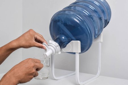Foto de La mano del hombre vierte agua en una taza de vidrio de un galón grande. Verter agua fresca y clara de un gran galón azul en un vaso - Imagen libre de derechos