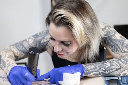 Foto horizontal: el enfoque detallado de una artista del tatuaje es palpable mientras trabaja meticulosamente en el tatuaje de un cliente, sus propios brazos entintados cuentan una historia de su dedicación. Concepto negocio, arte.