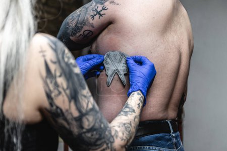 Fotografía horizontal primer plano de un artista del tatuaje aplicando una plantilla de transferencia de un diseño de escudo de armas en el cuerpo de un cliente. Concepto negocio, arte.