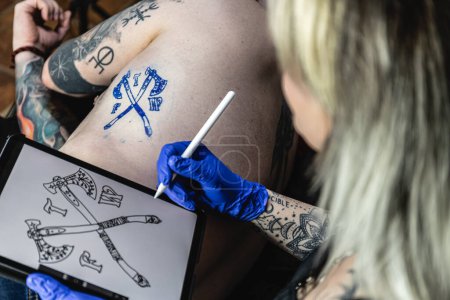 Fotografía horizontal un artista del tatuaje que comprueba el diseño de un escudo de armas en la piel de un cliente, haciendo referencia a un dibujo digital. Concepto negocio, arte.