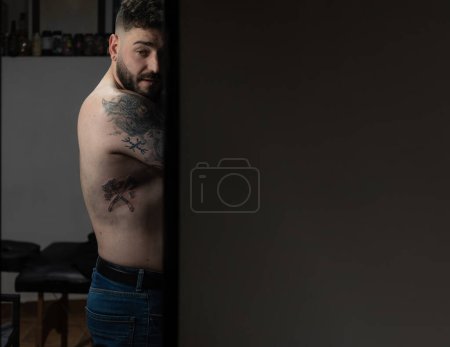 Horizontal photo a tattooed man looks back, his body art visible in a serene, dimly-lit tattoo studio, evoking a narrative of personal expression. Copy space. Concept business, art.