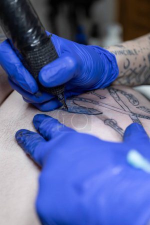 Photo verticale Une prise de vue ciblée capture les mains d'un tatoueur, vêtue de gants bleus, détaillant habilement un tatouage avec une machine à tatouer professionnelle. Concept business, art.
