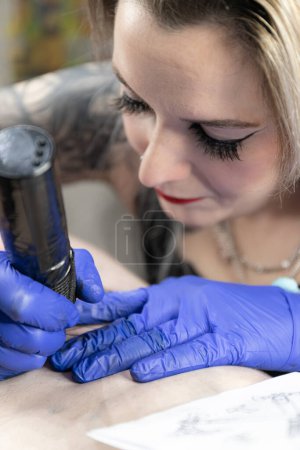 Vertikales Foto Eine intime Nahaufnahme einer Tätowiererin, während sie sich intensiv auf das Einfärben eines Designs konzentriert, wobei ihre blauen Handschuhe den Farbton des Tattoos hervorheben. Konzeptgeschäft, Kunst.