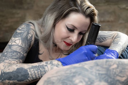 Horizontal photo a tattoo artist with full sleeve tattoos concentrates on inking a client, her expertise evident in her steady hand and focused gaze. Concept business, art.