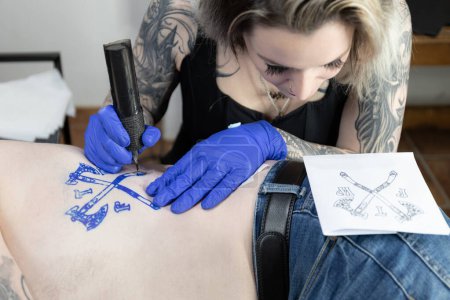 Horizontal photo tattoo artist meticulously inks a coat of arms design on the body, using a sketch as a guide for the intricate work. Concept business, art.