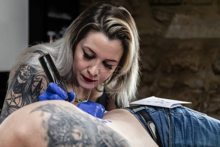 Foto horizontal: una artista del tatuaje enfocada trabaja diligentemente en un tatuaje de escudo de brazos, su atención al detalle evidente en su cuidadosa técnica. Concepto negocio, arte.